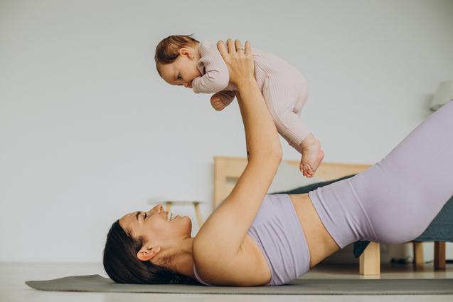 Mutter liegt auf einer Yogamatte und hebt fr&ouml;hlich ein Baby &uuml;ber sich, beides in sportlicher Kleidung.