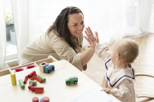 Frau spielt mit einem Kleinkind am Tisch, Holzspielzeug liegt verstreut auf der Tischplatte.