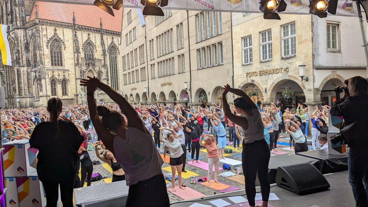 Gruppenyoga-Session im Freien mit Teilnehmern in einem Platz vor einer Kirche, umgeben von Gebäuden.