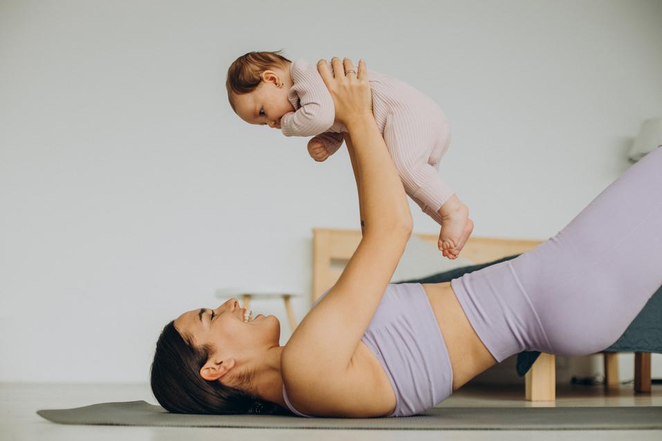 Baby Yoga Mutter liegt auf einer Yogamatte und hebt fröhlich ein Baby über sich, beides in sportlicher Kleidung.