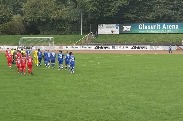 Zwei Fußballmannschaften in blauen und roten Trikots stehen auf einem grünen Rasenplatz in der Glasurit Arena.