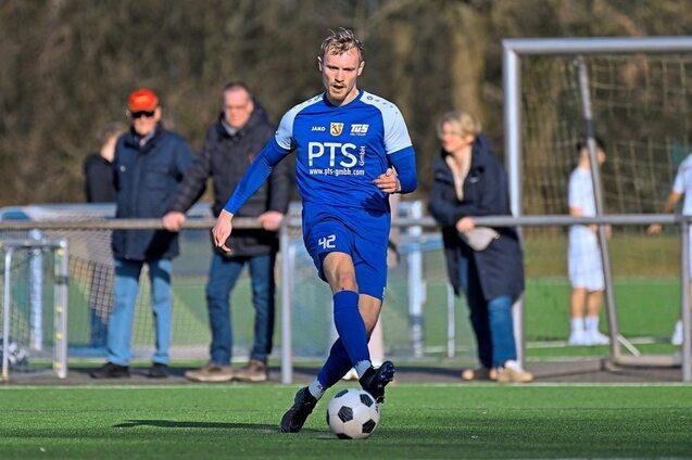 Ein Fu&szlig;ballspieler in blauer Uniform dribbelt mit einem Ball auf einem Kunstrasenplatz, Zuschauer im Hintergrund.
