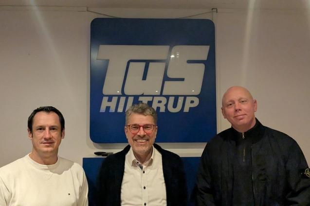 Drei Männer stehen vor einem Logo von TuS Hiltrup in einem Innenraum.