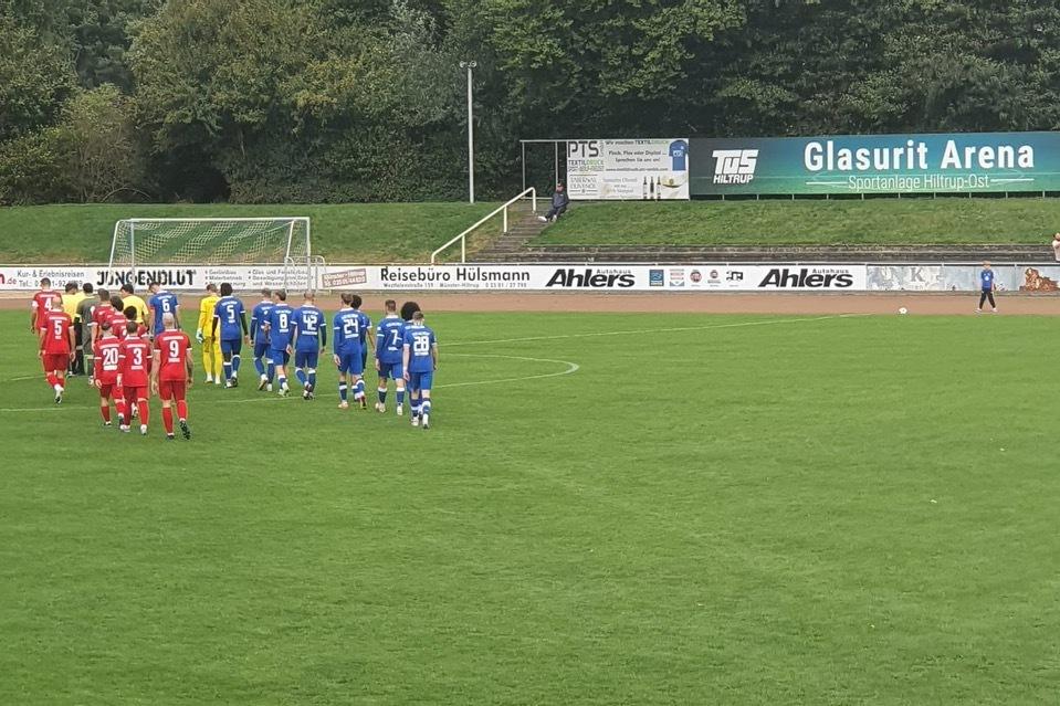 Zwei Fußballmannschaften in blauen und roten Trikots stehen auf einem grünen Rasenplatz in der Glasurit Arena.