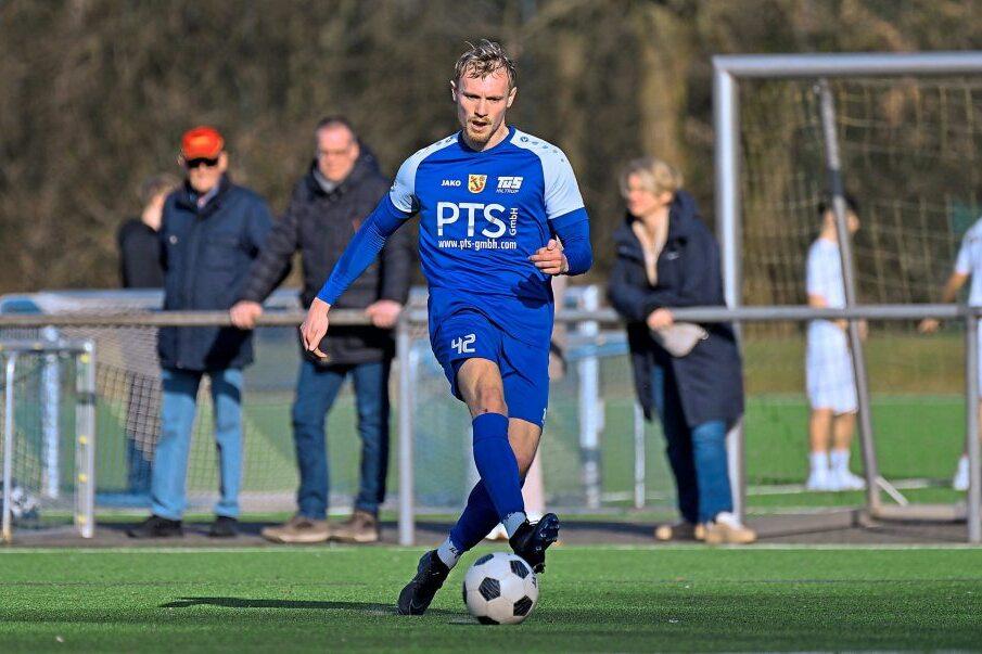 Ein Fu&szlig;ballspieler in blauer Uniform dribbelt mit einem Ball auf einem Kunstrasenplatz, Zuschauer im Hintergrund.