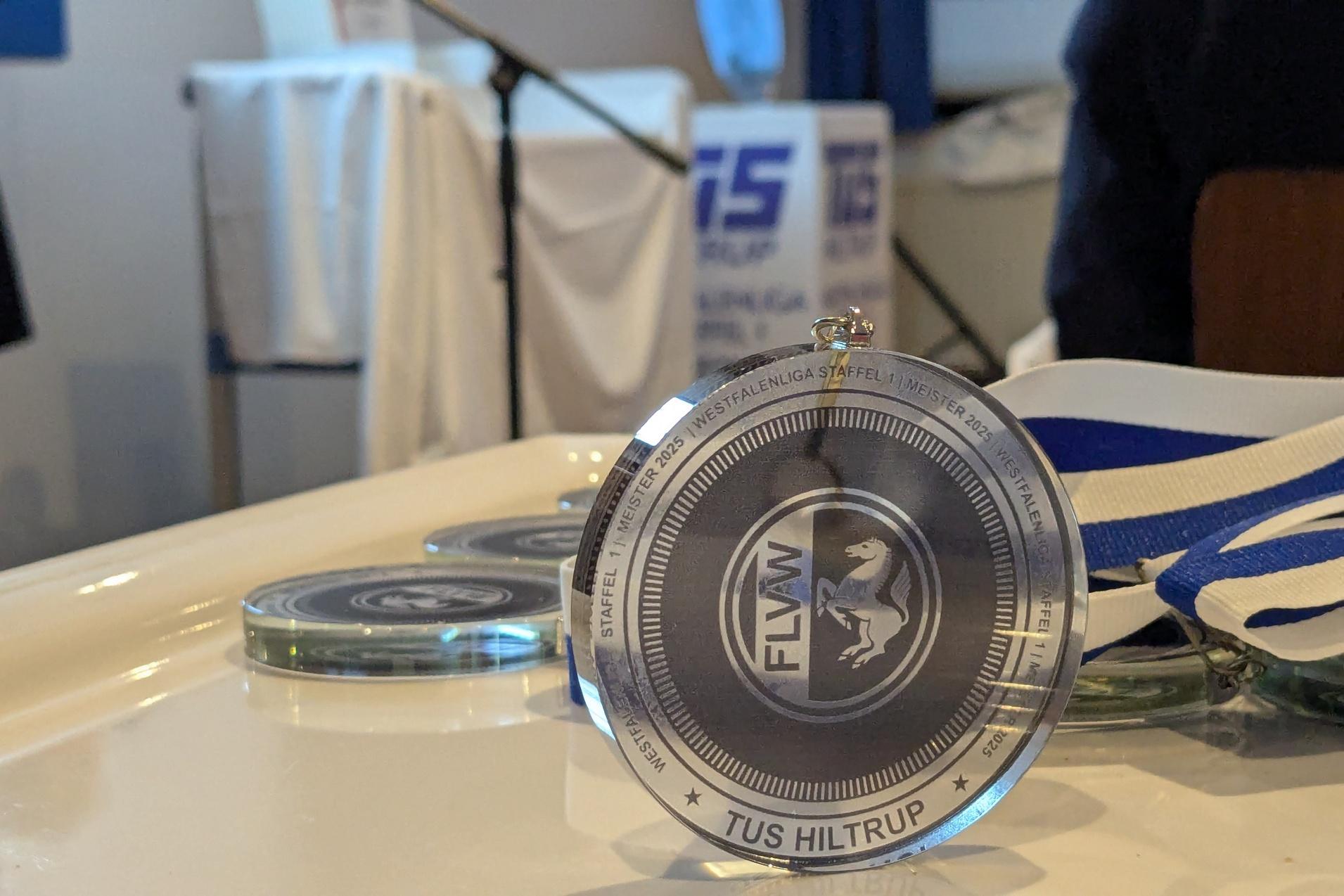 Nahaufnahme einer Siegermedaille mit der Gravur „TUS Hiltrup“ auf einem Tisch, umgeben von weiteren Medaillen.
