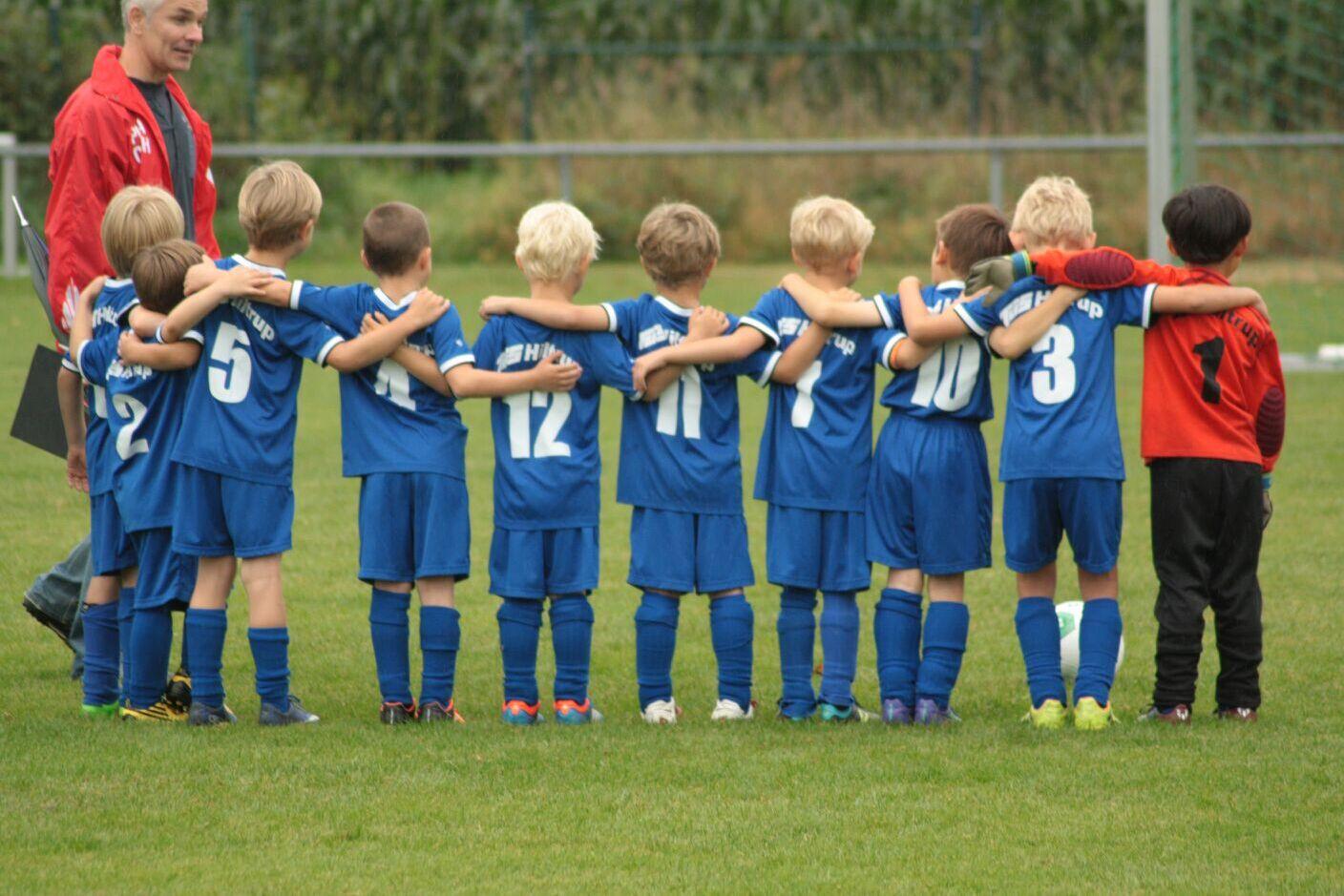 Gruppe von sieben Kindern in blauen Fu&szlig;balltrikots, die sich umarmen, w&auml;hrend ein Trainer daneben steht.