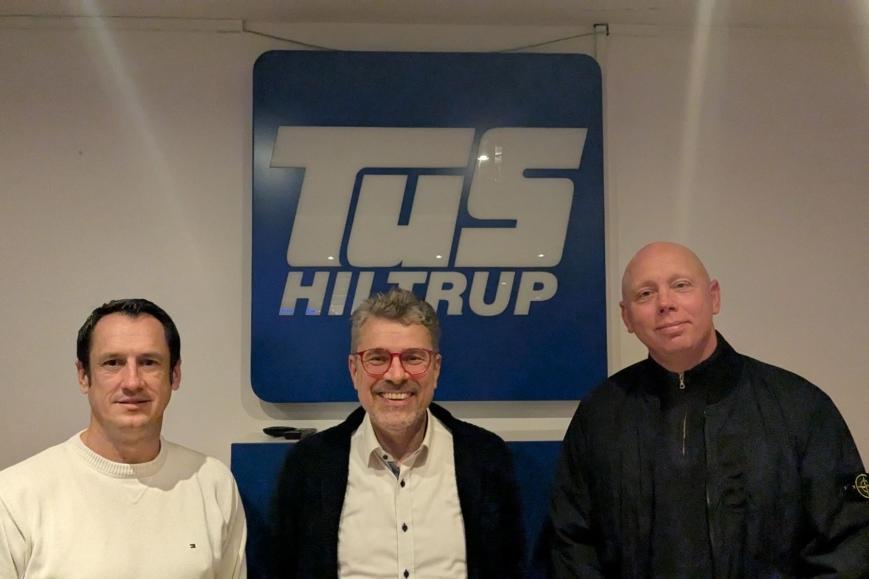Drei Männer stehen vor einem Logo von TuS Hiltrup in einem Innenraum.