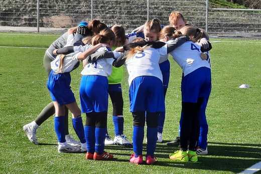 Gruppe von M&auml;dchen in Sportkleidung versammelt sich im Kreis auf einem Fu&szlig;ballplatz, um vor dem Spiel zu besprechen.