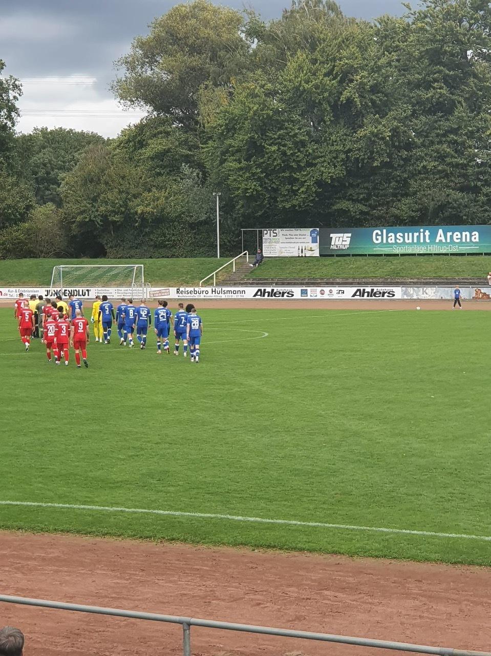 Zwei Fußballmannschaften in blauen und roten Trikots stehen auf einem grünen Rasenplatz in der Glasurit Arena.