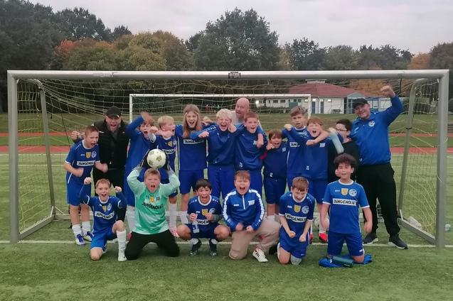 Gruppe von jubelnden jungen Fußballspielern in blauen Trikots, stehend vor einem Tor auf einem Sportplatz.