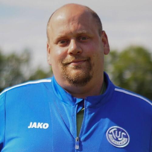 Ein Mann mit Glatze trägt eine blaue Trainingsjacke und lächelt vor verschwommenem Hintergrund.