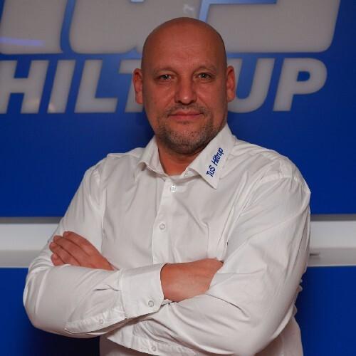 Mann mit kurzen Haaren in weißem Hemd, lächelt und hat die Arme vor der Brust verschränkt, vor blauer Wand.