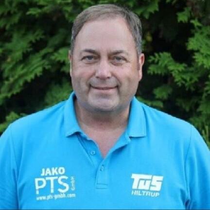 Mann in blauem Poloshirt mit Logos vor grünem Hintergrund lächelt in die Kamera.