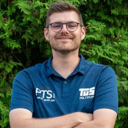 Mann mit Brille und verschränkten Armen in blauer Poloshirt vor grünem Hintergrund.