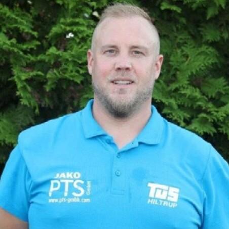 Mann mit kurzer Frisur trägt ein blaues Poloshirt mit Logodruck und steht vor grünem Hintergrund aus Bäumen.