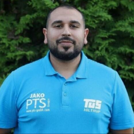 Porträt eines Mannes in blauem Poloshirt mit Logos, vor einem Hintergrund aus grünen Pflanzen.