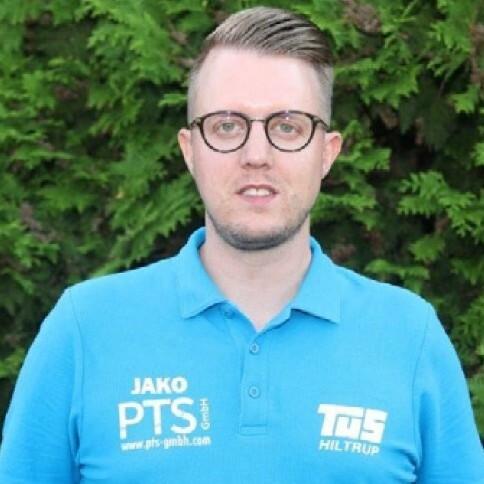 Mann mit Brille trägt ein blaues Poloshirt mit Logos vor grünem Hintergrund.