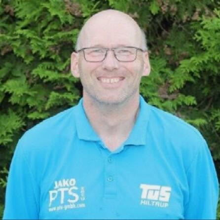 Portrait eines lächelnden Mannes mit Glatze in einem blauen Poloshirt vor einem grünen, bewachsenen Hintergrund.