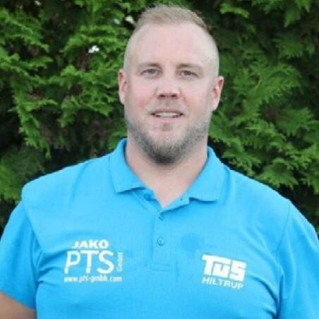 Mann mit kurzer Frisur trägt ein blaues Poloshirt mit Logodruck und steht vor grünem Hintergrund aus Bäumen.