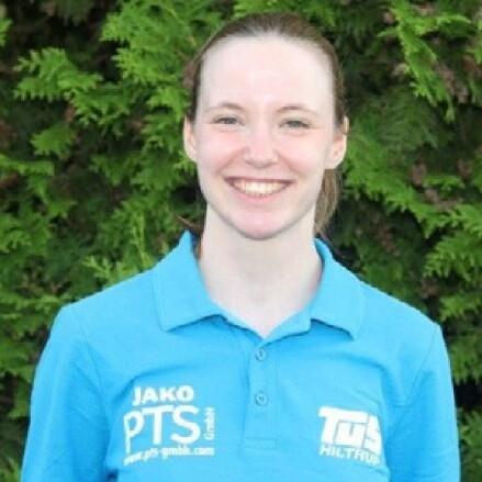 Lächelnde Frau mit langen Haaren, trägt ein blaues Poloshirt mit Logos auf grünem Hintergrund.