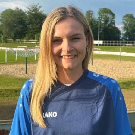 Portrait einer jungen Frau mit langen Haaren, die ein blaues Sporttrikot trägt, vor einem Fußballfeld im Hintergrund.