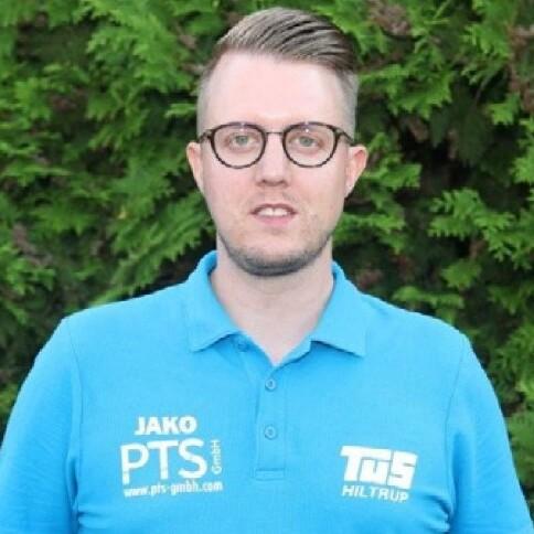 Mann mit Brille trägt ein blaues Poloshirt mit Logos vor grünem Hintergrund.