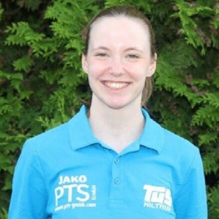 Lächelnde Frau mit langen Haaren, trägt ein blaues Poloshirt mit Logos auf grünem Hintergrund.