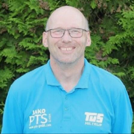 Portrait eines lächelnden Mannes mit Glatze in einem blauen Poloshirt vor einem grünen, bewachsenen Hintergrund.