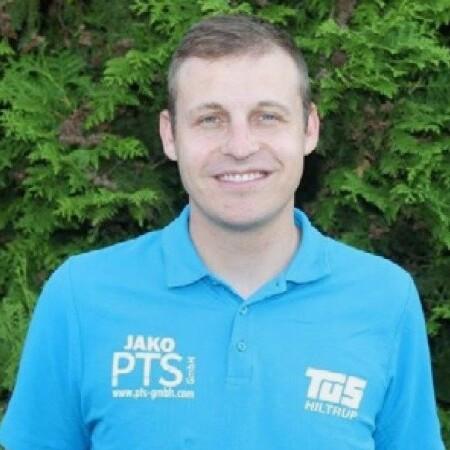 Portrait eines lächelnden Mannes in blauer Sportbekleidung vor grüner Pflanzenwand.