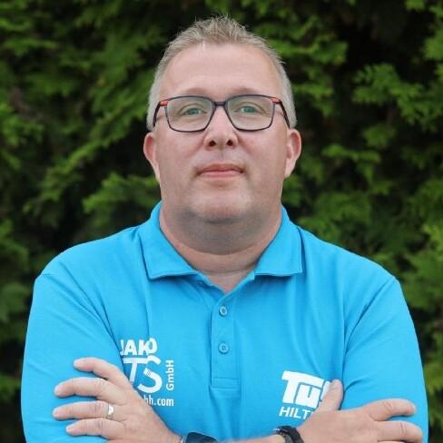 Porträt eines Mannes in blauer Polo-Shirt, mit Brille, verschränkt die Arme vor grünem Hintergrund.