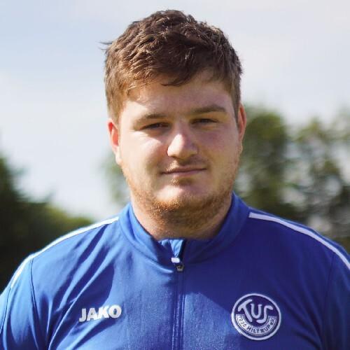 Porträt eines jungen Mannes in blauer Sportjacke mit Vereinslogo im Freien, unscharfer Hintergrund mit Bäumen.