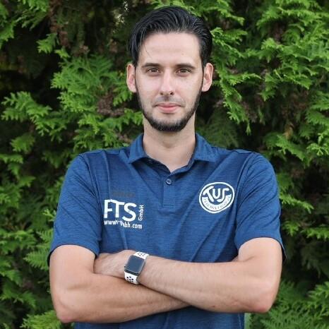 Mann in blauer Sportbekleidung steht vor grünen Büschen mit verschränkten Armen und neutralem Ausdruck.