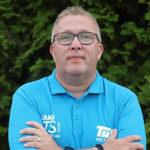 Porträt eines Mannes in blauer Polo-Shirt, mit Brille, verschränkt die Arme vor grünem Hintergrund.
