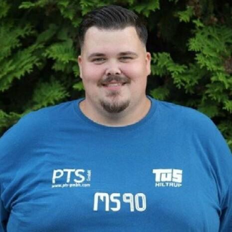 Mann mit Kurzhaarfrisur trägt ein blaues T-Shirt vor grünem Hintergrund, lächelt direkt in die Kamera.