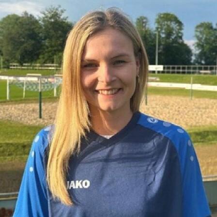 Portrait einer jungen Frau mit langen Haaren, die ein blaues Sporttrikot trägt, vor einem Fußballfeld im Hintergrund.