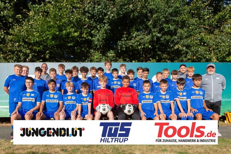 Teamfoto einer Jugendfußballmannschaft in blauen Trikots, umrahmt von Sponsorenlogos, im Freien auf einer Sportanlage.