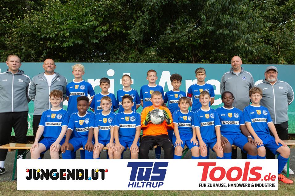 Teambild einer Jugendfußballmannschaft in blauen Trikots, umgeben von zwei Trainern vor einer grünen Wand.