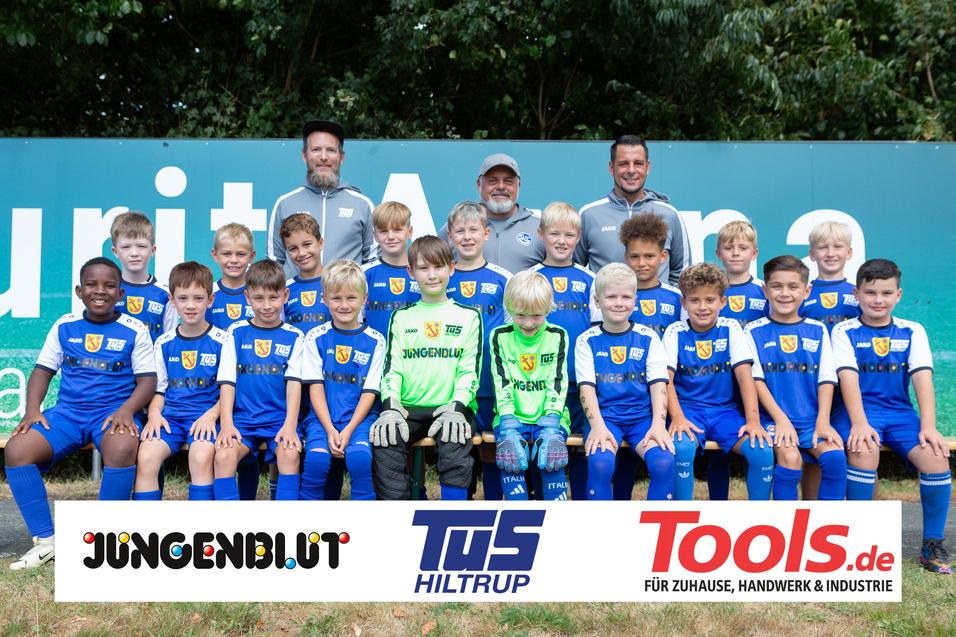 Gruppenfoto einer Jugendfußballmannschaft mit drei Trainern, alle in blauen Trikots, vor einem Werbebanner.