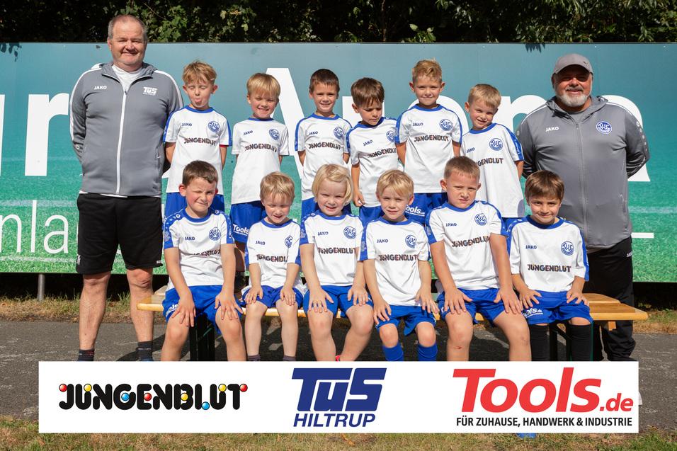 Gruppenfoto einer Jugendfußballmannschaft mit Trainer im Sportoutfit vor einer Werbetafel und auf einer Wiese.
