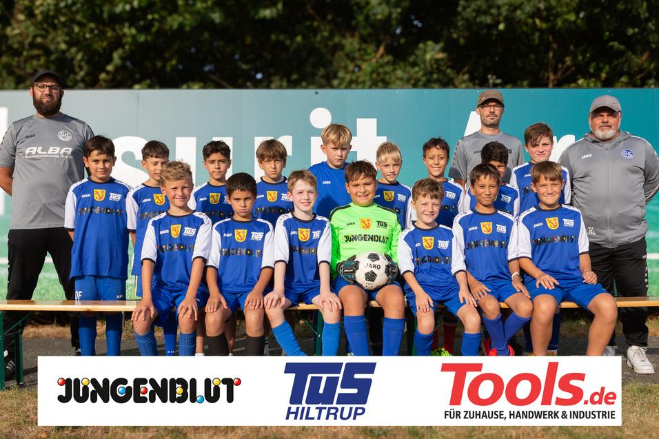 Gruppenfoto einer Jugendfußballmannschaft mit dem Trainer, in blauen Trikots auf einer Wiese neben einer Werbetafel.