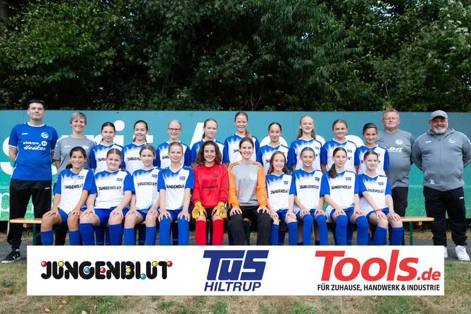 Mannschaftsfoto einer Mädchenfußballmannschaft in blauen Trikots, umgeben von Trainern und Sponsorenlogos.