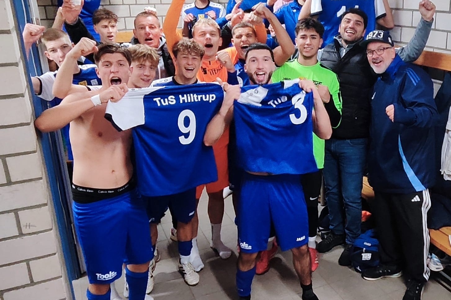 Jubilierende Fußballmannschaft von TuS Hiltrup feiert im Umkleideraum, trägt blaue Trikots und zeigt stolz ihre Nummern.