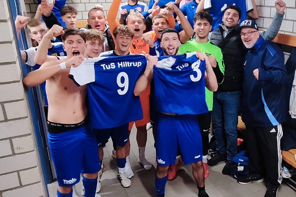 Jubilierende Fußballmannschaft von TuS Hiltrup feiert im Umkleideraum, trägt blaue Trikots und zeigt stolz ihre Nummern.