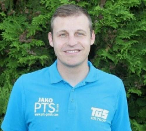 Mann mit kurzem, braunem Haar trägt ein blaues Poloshirt und lächelt vor einer grünen, bunten Hintergrundpflanze.