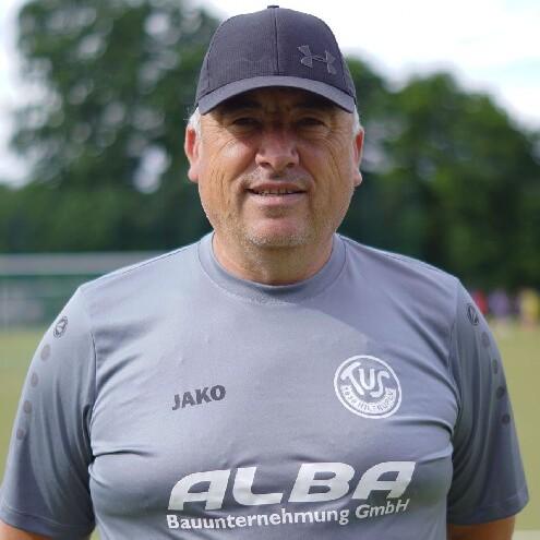 Mann mit Cap steht l&auml;chelnd vor einem Fu&szlig;ballplatz, tr&auml;gt graues Teamtrikot mit Logo und Sponsorenaufdruck.