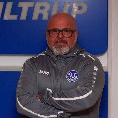 Mann mit Glatze und Brille in grauer Sportjacke, mit verschränkten Armen vor blauem Hintergrund.