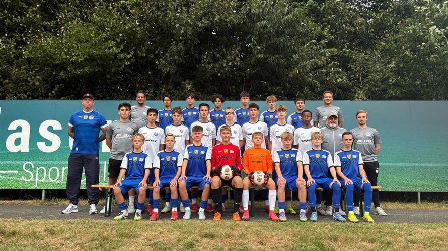 Gruppenfoto einer Fußballmannschaft mit Spielern in blauen und weißen Trikots, umgeben von Trainern und einem Werbeschild.