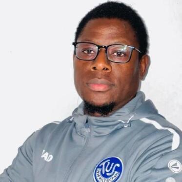 Porträt eines Mannes mit Brille in grauem Sportanzug, der mit verschränkten Armen in die Kamera schaut.