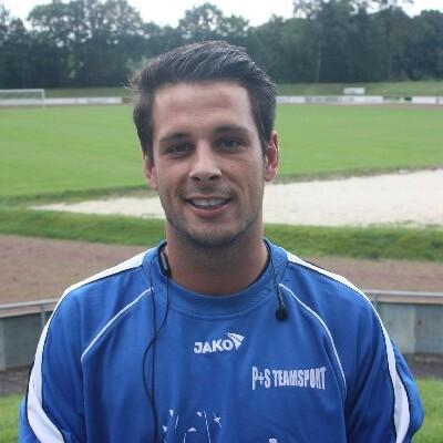 Portrait eines lächelnden Mannes in Sportkleidung, stehend auf einem Fußballplatz mit grünem Rasen im Hintergrund.